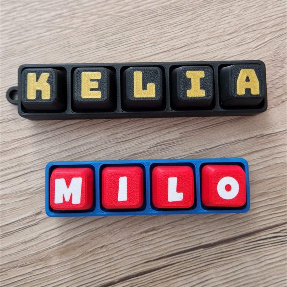 Deux plaques prénoms imprimées en 3D à lettres amovibles : KELIA en noir et or, MILO en bleu et rouge.